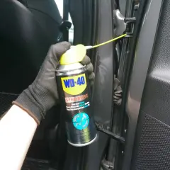 Ręka w rękawiczce aplikuje biały smar litowy WD-40 na zawiasy drzwi, rozwiązując problem czym smarować zawiasy w drzwiach.