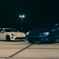 Biały coupe i niebieski hatchback stoją nocą na parkingu.