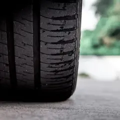 Widoczne wybrzuszenie na zużytej oponie, która dotyka asfaltu. W tle zamazana zieleń drzew.
