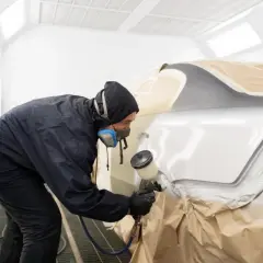 Lakiernik w masce i kombinezonie maluje samochód w kabinie. Czas schnięcia lakieru samochodowego zależy od wielu czynników.