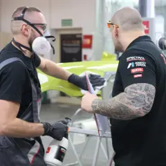 Dwóch mężczyzn w maskach i rękawiczkach przygotowuje element karoserii. Czy podkład epoksydowy trzeba matowić? To pytanie zadają sobie podczas pracy.