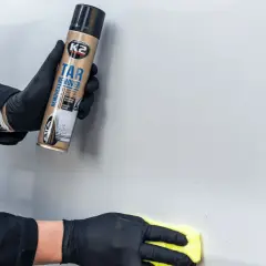 Ręce w czarnych rękawiczkach używają preparatu K2 Tar Remover, by usunąć naklejki z samochodu. Żółta ściereczka poleruje powierzchnię.