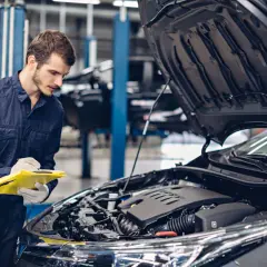 Mechanik w granatowym kombinezonie sprawdza listę kontrolną przy otwartej masce samochodu. Konie mechaniczne skrót do szybkiej diagnozy.