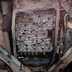 Widok od spodu na otwartą skrzynię automatyczną, gotową do wymiany oleju. Widać skomplikowany mechanizm i ślady zużytego płynu.