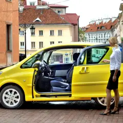 Żółte, dziwne samochody osobowe na brukowanej uliczce. Kobieta w białej koszuli i czarnej spódnicy stoi obok jednego z nich.