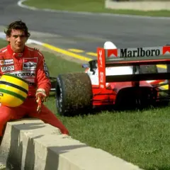 Ayrton Senna w czerwonym kombinezonie siedzi na krawężniku, trzymając swój żółty kask. Za nim stoi czerwony bolid Formuły 1. To chwila przed tragicznym wypadkiem.