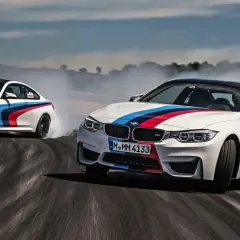 Dwa białe samochody do driftu z kolorowymi pasami M BMW na torze, unoszą kłęby dymu.