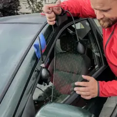 Mężczyzna w czerwonej kurtce wykonuje otwieranie awaryjne samochodu, używając narzędzia do podważenia drzwi.