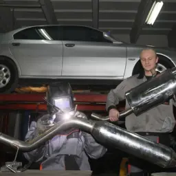 Mechanik spawa nowy wydech w samochodzie. Obok stoi kolega z gotowym tłumikiem.