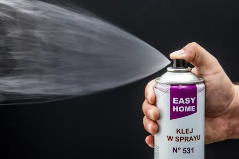 Dłoń trzyma puszkę z napisem "EASY HOME KLEJ W SPRAYU", rozpylając białą mgiełkę. Idealny do podsufitki.