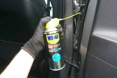 Ręka w rękawiczce aplikuje biały smar litowy WD-40 na zawiasy drzwi, rozwiązując problem czym smarować zawiasy w drzwiach.