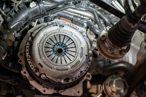 Mechanik pracuje nad grawitacyjnym odpowietrzaniem sprzęgła w samochodzie. Widoczny jest mechanizm sprzęgła i elementy silnika.