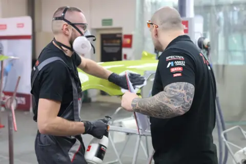 Dwóch mężczyzn w maskach i rękawiczkach przygotowuje element karoserii. Czy podkład epoksydowy trzeba matowić? To pytanie zadają sobie podczas pracy.