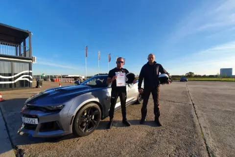 Dwóch mężczyzn z kaskami i certyfikatem po udanym kursie jazdy ekstremalnej obok sportowego samochodu.