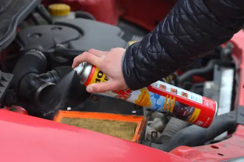 Ręka trzyma puszkę z preparatem samostart do diesla, rozpylając go w pobliżu filtra powietrza w silniku samochodu.