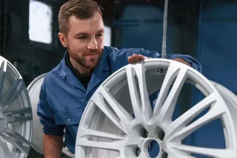 Pracownik w niebieskim kombinezonie sprawdza stan felgi aluminiowej, przygotowując ją do renowacji. Proces jak odnowić alufelgi wymaga precyzji.