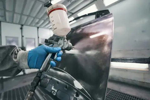 Ręka w niebieskiej rękawiczce trzyma pistolet lakierniczy, przygotowując element do malowania. Pytanie, czy na podkład epoksydowy można kłaść lakier, jest kluczowe dla tego procesu.