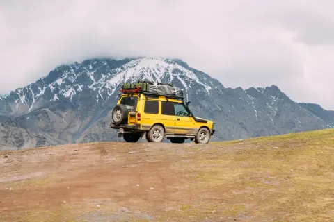 Żółty off road 4x4 z bagażem na dachu i zapasowym kołem na tyle, zaparkowany na zboczu góry, pod ośnieżonymi szczytami.