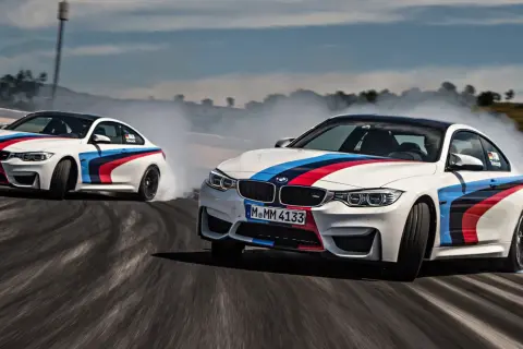 Dwa białe samochody do driftu z kolorowymi pasami M BMW na torze, unoszą kłęby dymu.
