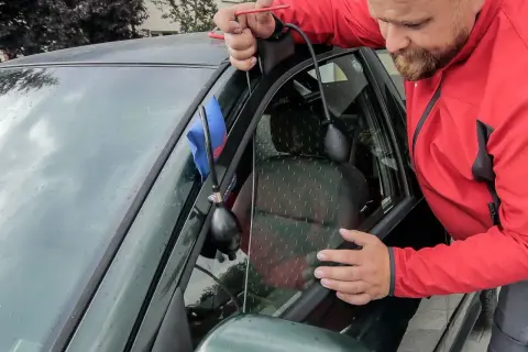 Mężczyzna w czerwonej kurtce wykonuje otwieranie awaryjne samochodu, używając narzędzia do podważenia drzwi.