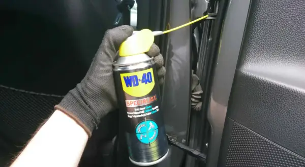 Ręka w rękawiczce aplikuje biały smar litowy WD-40 na zawiasy drzwi, rozwiązując problem czym smarować zawiasy w drzwiach.