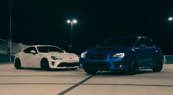 Biały coupe i niebieski hatchback stoją nocą na parkingu.