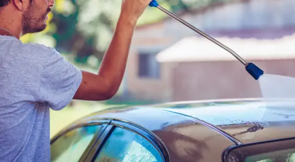 Mężczyzna pokazuje, jak myć auto na myjni, używając myjki ciśnieniowej do spłukiwania dachu samochodu.