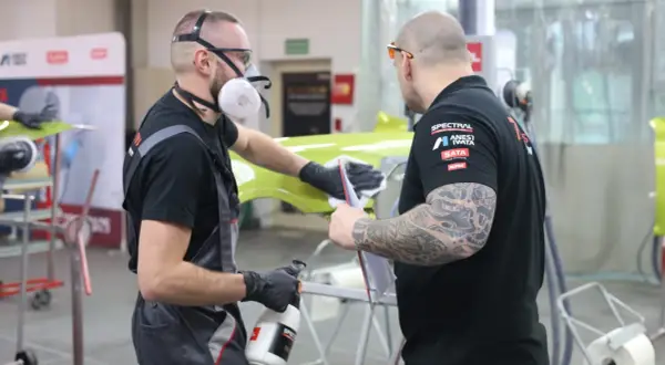 Dwóch mężczyzn w maskach i rękawiczkach przygotowuje element karoserii. Czy podkład epoksydowy trzeba matowić? To pytanie zadają sobie podczas pracy.