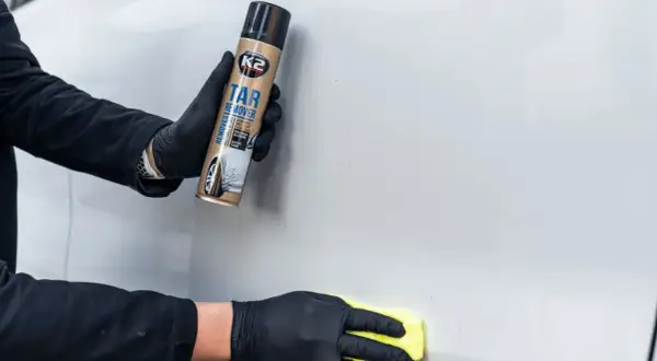 Ręce w czarnych rękawiczkach używają preparatu K2 Tar Remover, by usunąć naklejki z samochodu. Żółta ściereczka poleruje powierzchnię.