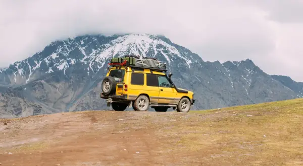 Żółty off road 4x4 z bagażem na dachu i zapasowym kołem na tyle, zaparkowany na zboczu góry, pod ośnieżonymi szczytami.