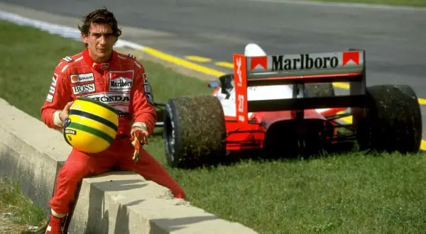 Ayrton Senna w czerwonym kombinezonie siedzi na krawężniku, trzymając swój żółty kask. Za nim stoi czerwony bolid Formuły 1. To chwila przed tragicznym wypadkiem.