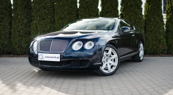 Elegancki, ciemnogranatowy Bentley Continental GT, brytyjski producent luksusowych aut, zaparkowany przed żywopłotem.