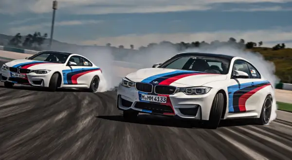 Dwa białe samochody do driftu z kolorowymi pasami M BMW na torze, unoszą kłęby dymu.