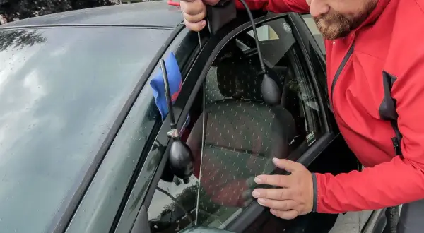 Mężczyzna w czerwonej kurtce wykonuje otwieranie awaryjne samochodu, używając narzędzia do podważenia drzwi.