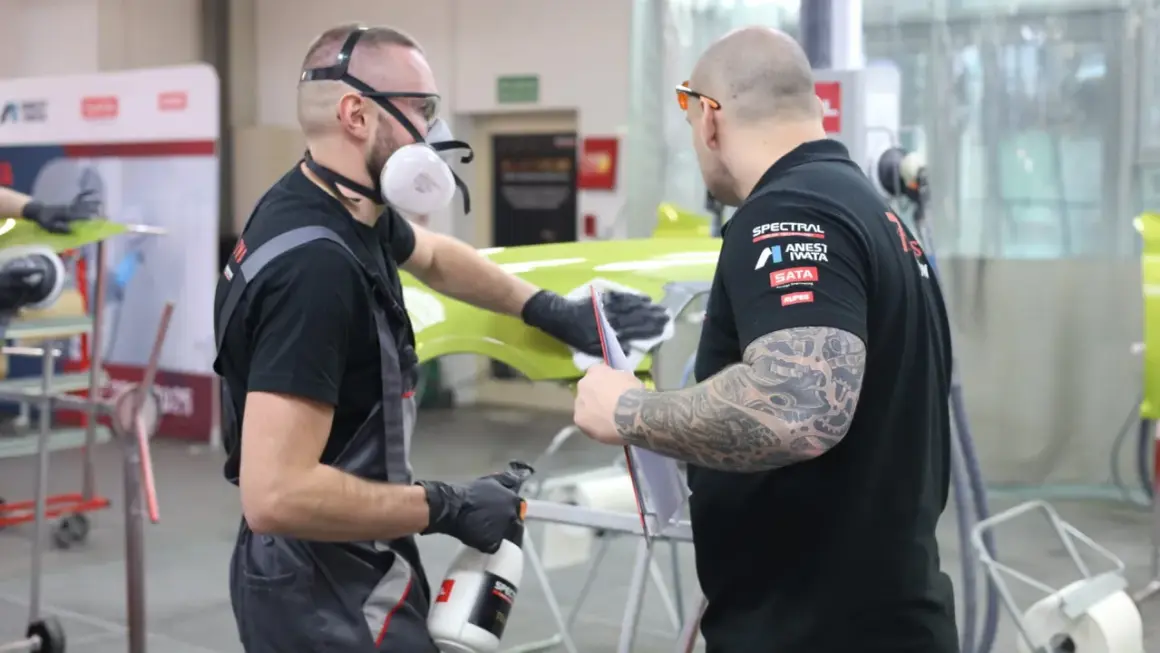Dwóch mężczyzn w maskach i rękawiczkach przygotowuje element karoserii. Czy podkład epoksydowy trzeba matowić? To pytanie zadają sobie podczas pracy.
