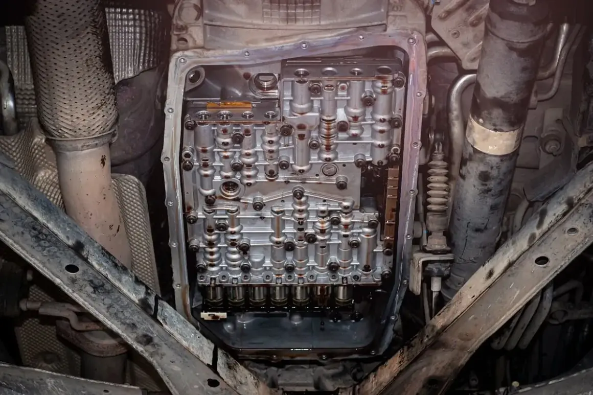 Widok od spodu na otwartą skrzynię automatyczną, gotową do wymiany oleju. Widać skomplikowany mechanizm i ślady zużytego płynu.
