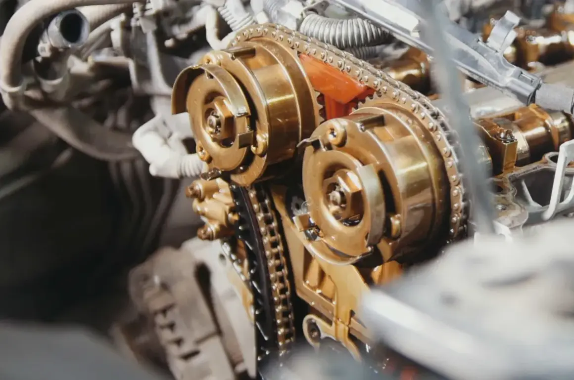 Zbliżenie na mechanizm silnika, gdzie trwa wymiana łańcucha rozrządu. Widoczne koła zębate i metalowy łańcuch pokryte olejem.