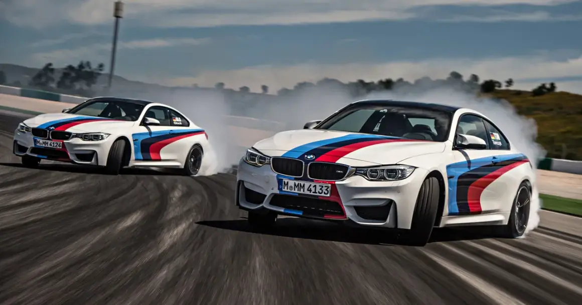 Dwa białe samochody do driftu z kolorowymi pasami M BMW na torze, unoszą kłęby dymu.