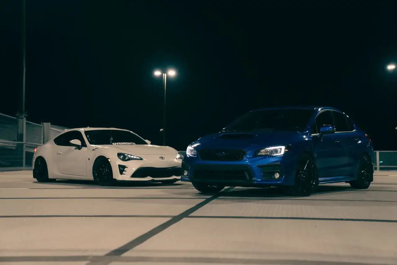 Biały coupe i niebieski hatchback stoją na parkingu nocą.