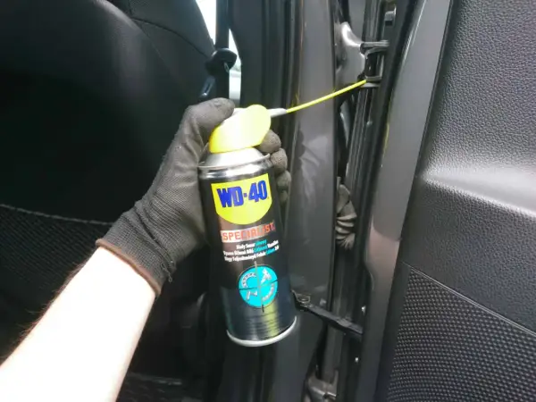 Ręka w rękawiczce aplikuje biały smar litowy WD-40 na zawiasy drzwi, rozwiązując problem czym smarować zawiasy w drzwiach.