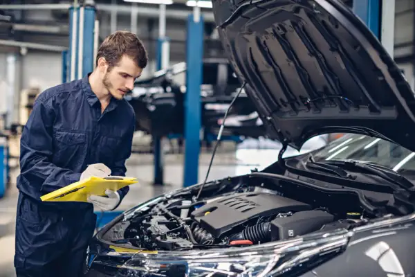 Mechanik w granatowym kombinezonie sprawdza listę kontrolną przy otwartej masce samochodu. Konie mechaniczne skrót do szybkiej diagnozy.